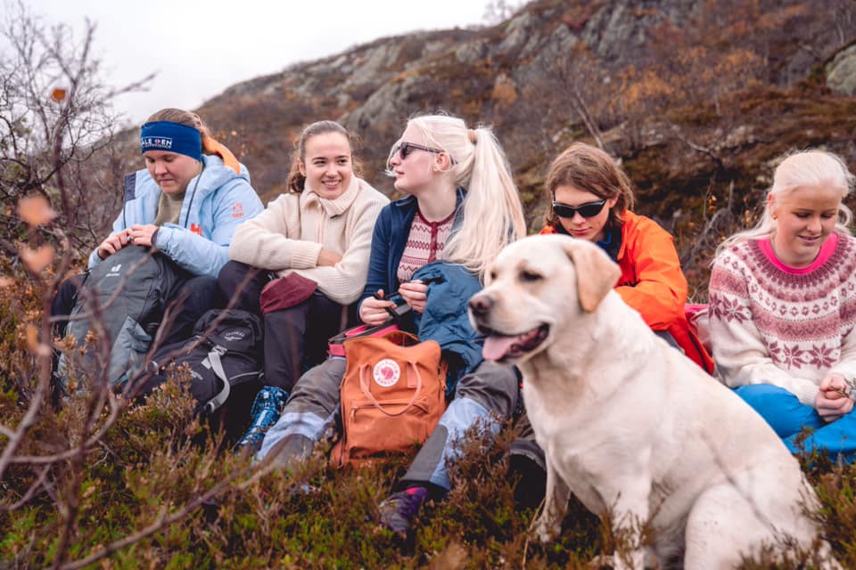 Fem medlemmer fra NBfU sitter på en fjelltopp og snakker sammen. Det er høst, og alle har på seg ullegensere. I tillegg ser vi en hvit førerhund på bildet, som sitter å slapper av. Alle smiler. 